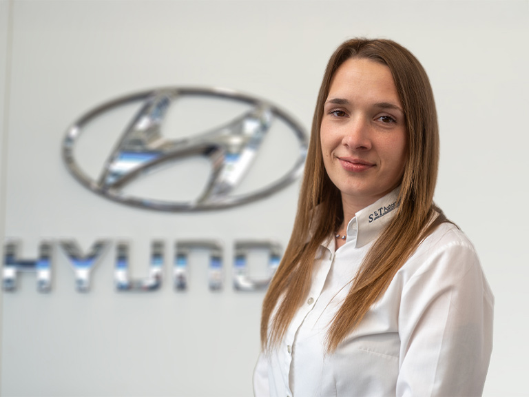 Auf diesem Foto ist die Serviceassistentin Nadja Stein vor einer hellgrauen Wand mit dem Logo von Hyundai.